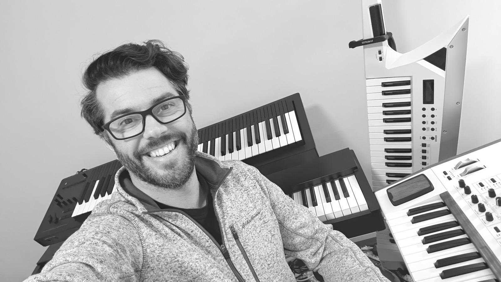 Aaron Hnatiw smiling in his studio, surrounded by keyboards, including a keytar.